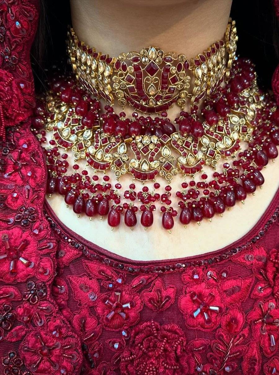 Macro of a layered ruby and gold bridal necklace over a deep red embroidered neckline, kundan stones and ruby drops stacked in tiers